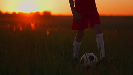 Close-up-of-Tracking-the-feet-of-a-football-boy-in-a-red-t-shirt-and-shorts-running-with-the-ball-at-sunset-in-the-field-on-the-grass.-The-young-football-player-dreams-of-a-professional-career-and-trains-in-the-field.-On-the-way-to-success